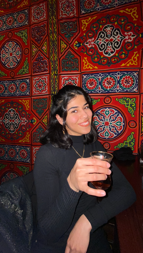 Ola holding a cup of tea in front of a colorful tapestry